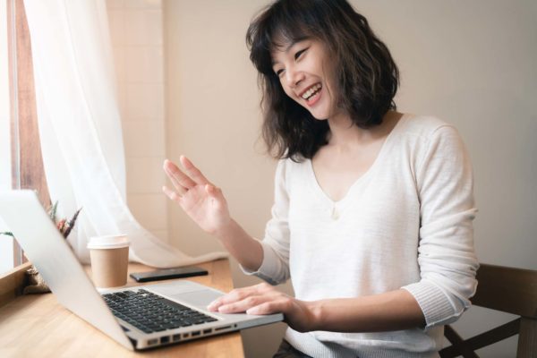 Young Asian woman working and video conferencing with laptop computer. A happy woman with smiley face working from home. A cup of coffee on table. Work from home concept.